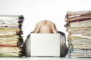 Man with his head in his hands, behind a laptop, with stacks of paper on either side of him. Clearly overwhelmed by volume of work!